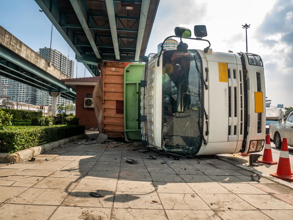 An overturned truck transporting a container on a road beneath a bridge at an intersection.